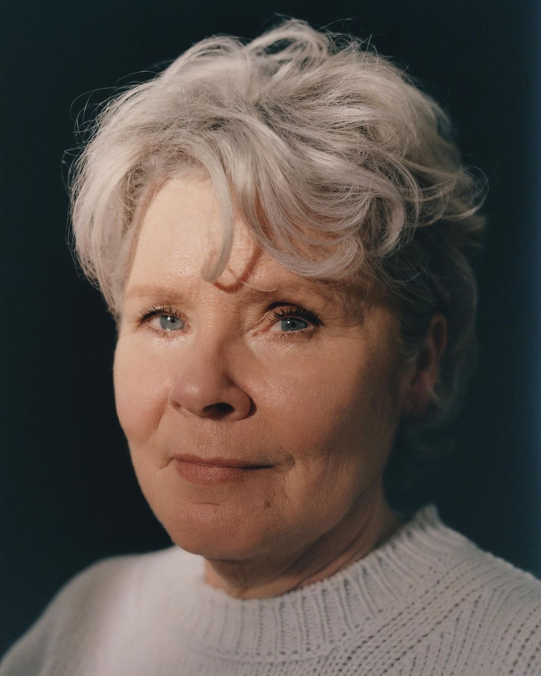 Headshot of an elderly woman with gray hair and a white sweater against a dark background, captured with soft, diffused studio lighting.