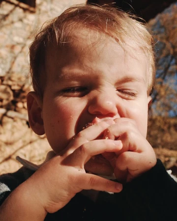Close-up of a young child with blond hair eating, holding food in their hands with a blurred outdoor background featuring trees.