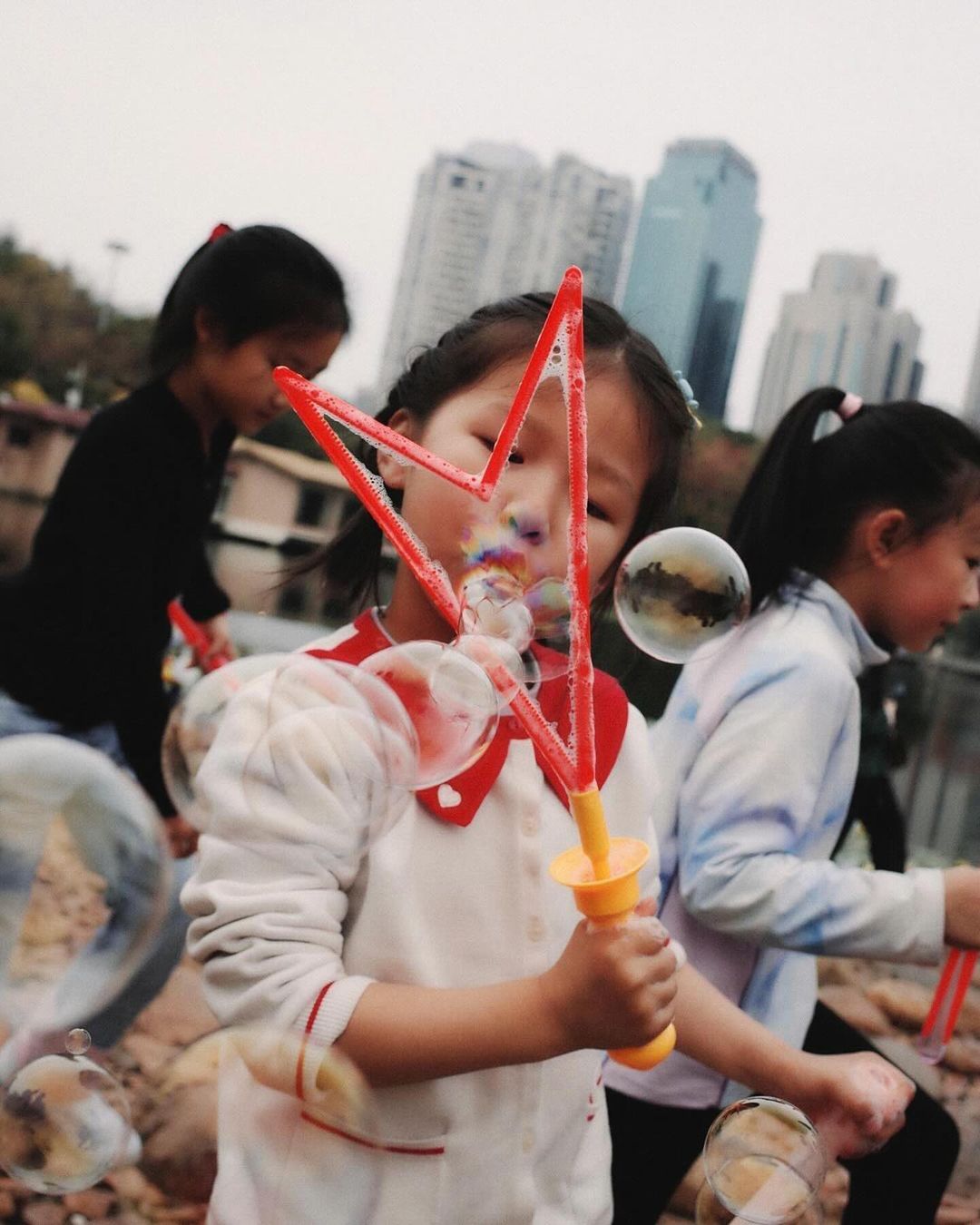 Three young Asian girls enjoy making bubbles outdoors near buildings, wearing casual clothing on a cloudy day, creating a joyful scene.