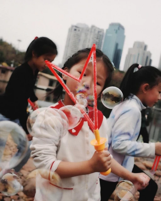 Three young Asian girls enjoy making bubbles outdoors near buildings, wearing casual clothing on a cloudy day, creating a joyful scene.