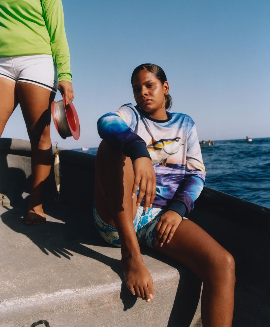 Two women on a boat at sea. One is sitting while the other is standing and holding fishing line. Clear blue skies and open ocean.