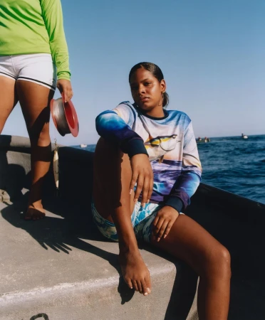 Two women on a boat at sea. One is sitting while the other is standing and holding fishing line. Clear blue skies and open ocean.