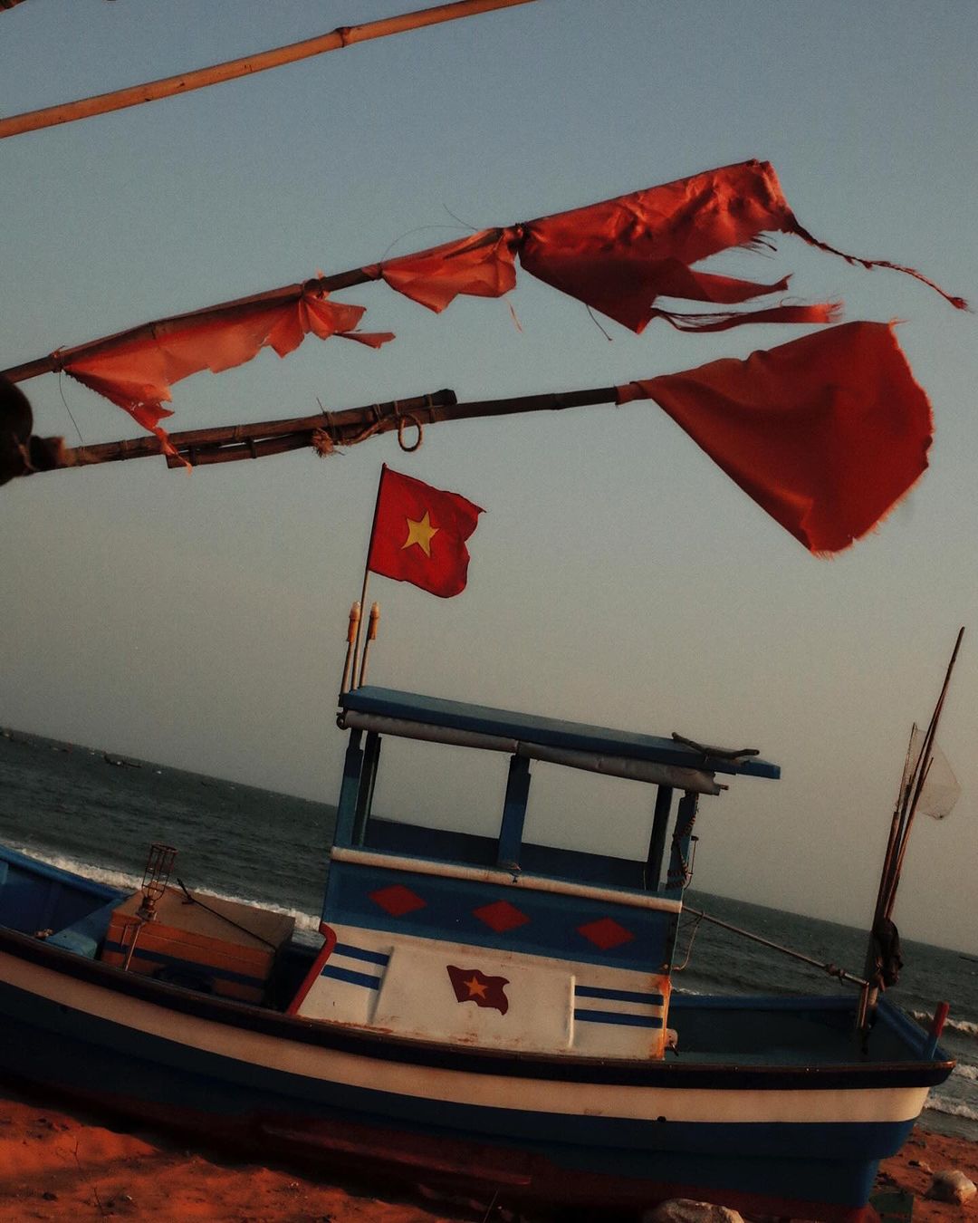 A blue and white boat with a red flag sits on a sandy beach by the ocean under a clear sky. Tattered red flags wave above.
