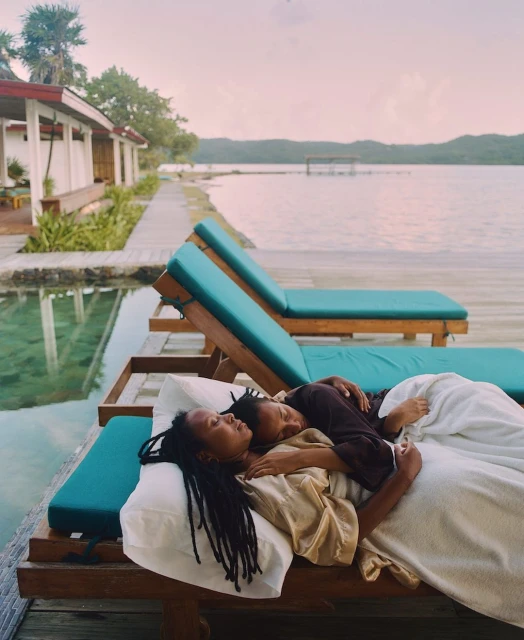Two women relax on lounge chairs by a pool overlooking a lake under a pastel sky. The scene evokes a peaceful vacation ambiance.