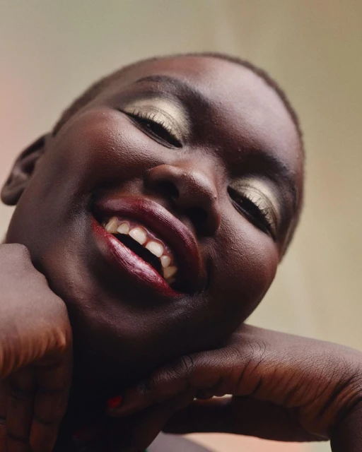 Close-up of a dark-skinned woman smiling with golden eyeshadow and dark red lipstick against a neutral background. Head and shoulders.
