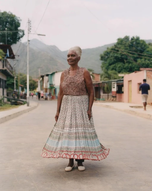 A medium shot shows an elderly woman with grey hair in a patterned dress, standing on a road in a residential area with mountains in the background.