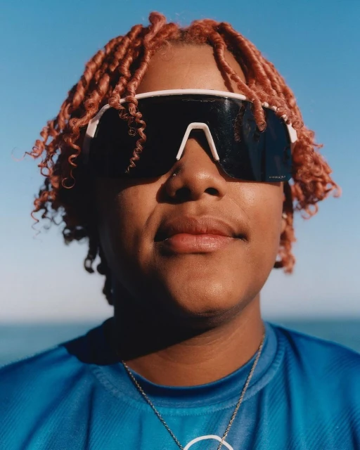 Close-up portrait of a person with pink braided hair wearing sunglasses and a blue athletic shirt with ocean and sky background.