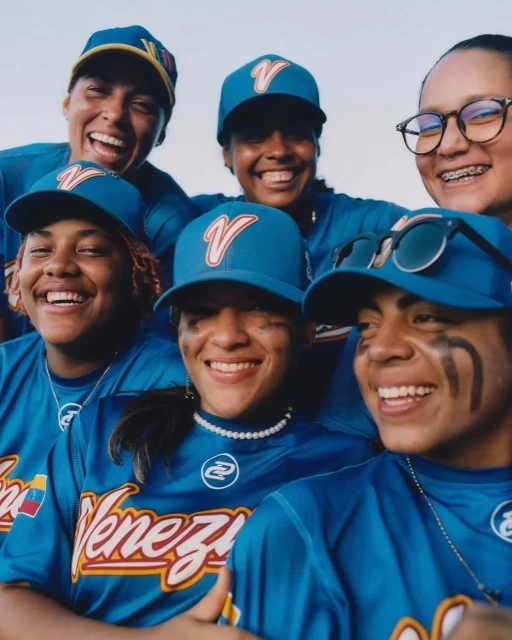 Six smiling young baseball players in blue uniforms and caps gather for a team portrait against a bright, neutral background.