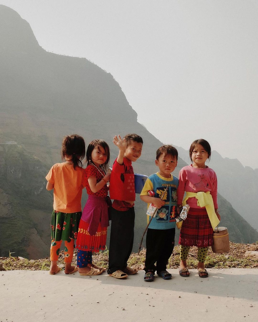Five children in brightly colored clothing stand in a row outdoors, smiling and posing in front of a hazy mountain backdrop.
