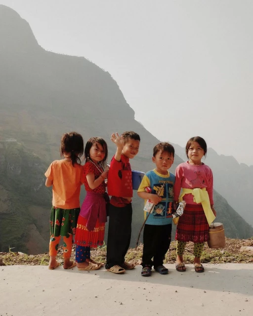 Five children in brightly colored clothing stand in a row outdoors, smiling and posing in front of a hazy mountain backdrop.