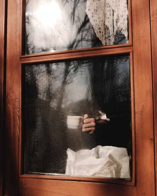 Person holding a coffee mug near a window with a view of trees. A white crocheted curtain and crumpled paper are visible inside.