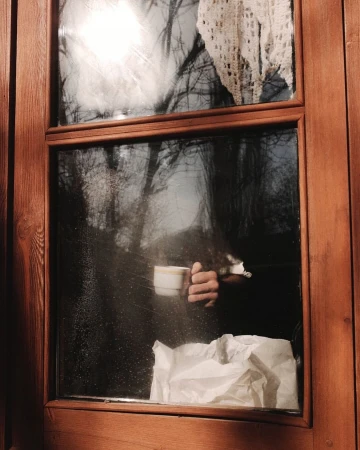 Person holding a coffee mug near a window with a view of trees. A white crocheted curtain and crumpled paper are visible inside.
