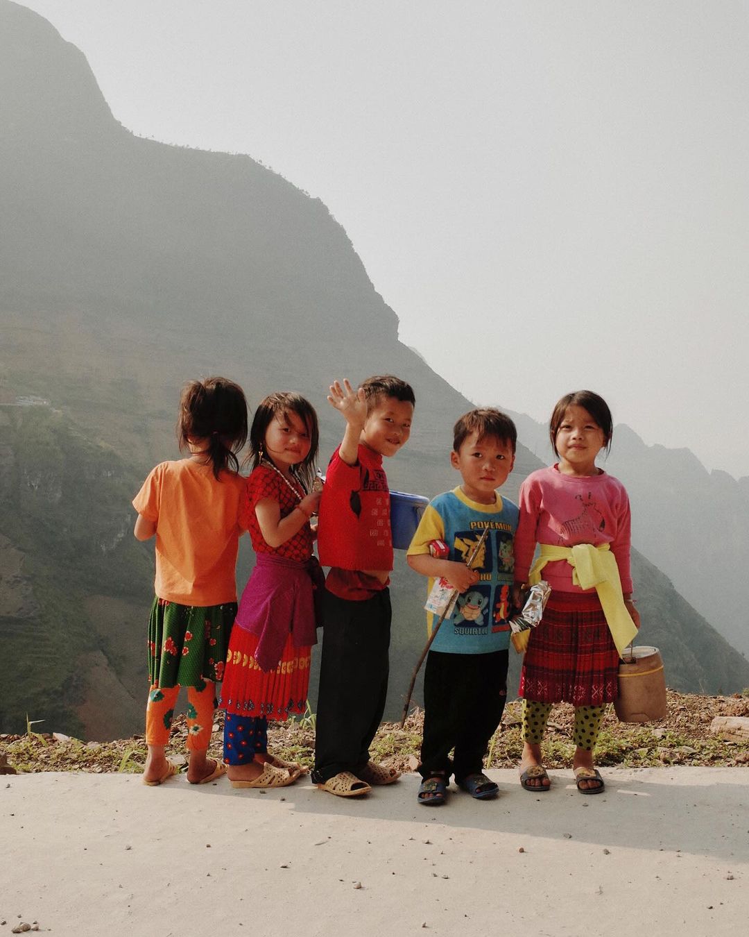 A group of five children of Asian descent stand together on a path with a mountain range visible in the background on a bright day.
