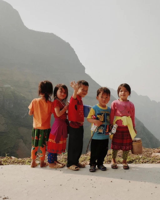 A group of five children of Asian descent stand together on a path with a mountain range visible in the background on a bright day.