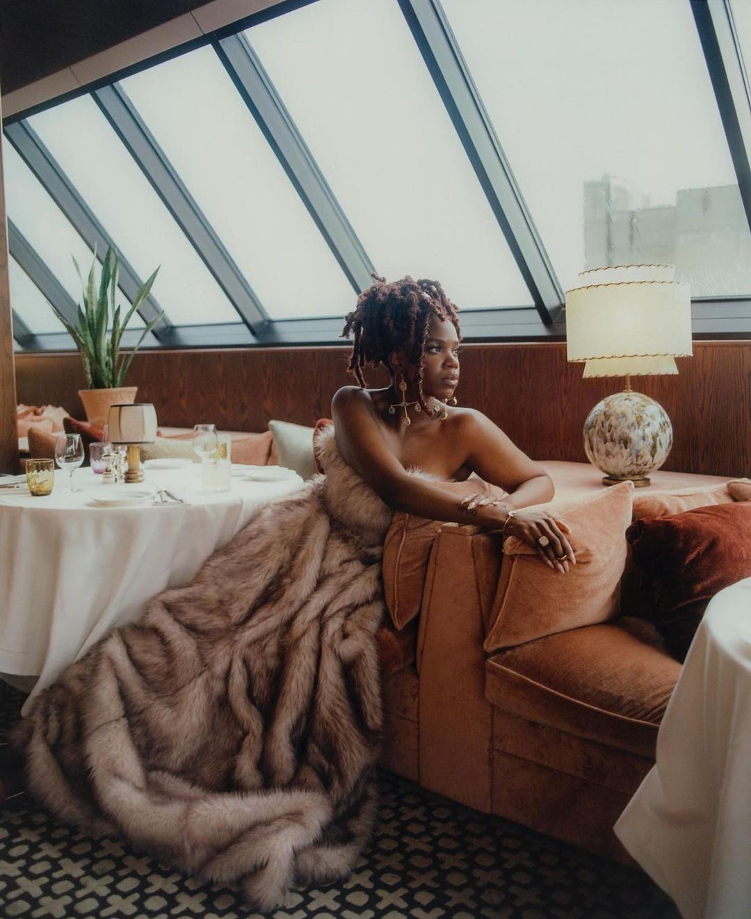Elegant woman in a fur-like wrap sits on a couch in a restaurant with modern architecture and neutral tones, beside a set table.