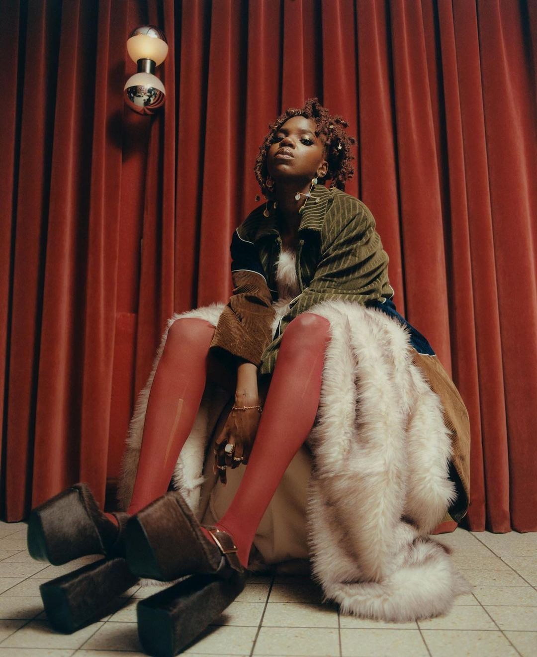 A stylish Black woman sits in chunky platform shoes and red tights, accented by fur coat and green jacket, with a red curtain backdrop.