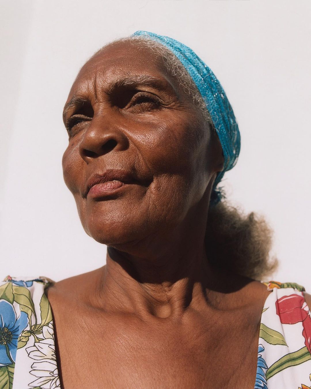 Close-up portrait of an elderly Black woman with a blue headscarf, wearing a floral top against a bright white background.