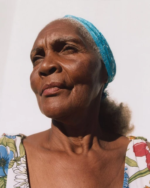 Close-up portrait of an elderly Black woman with a blue headscarf, wearing a floral top against a bright white background.