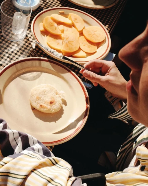 Overhead shot of persimmon slices and a small baked good on plates. Person holds a fork. Striped shirt and checkered tablecloth visible.