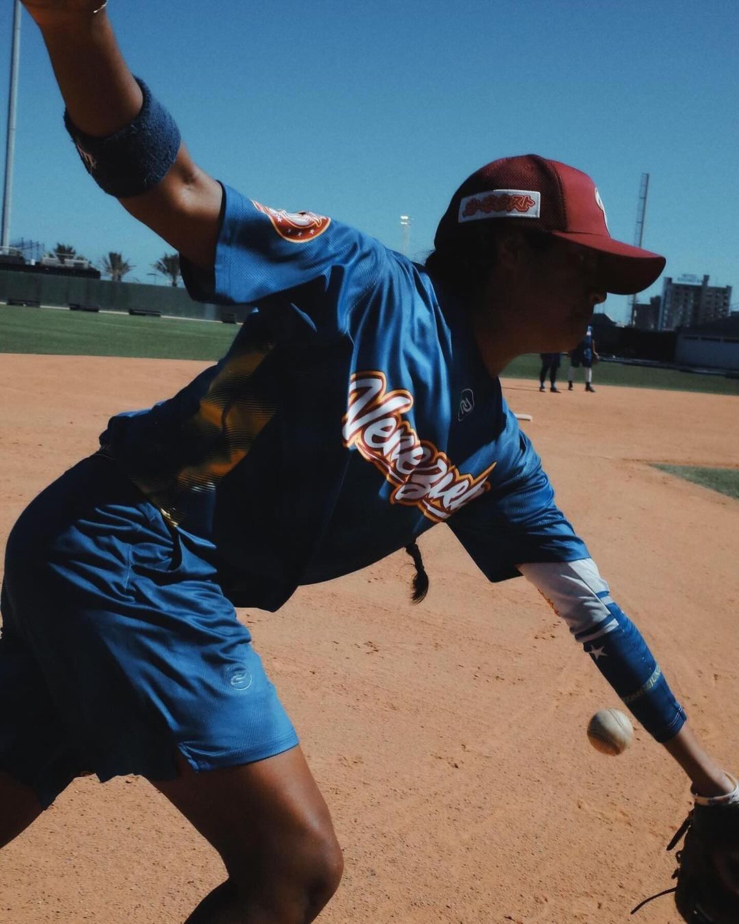 Female baseball player in action on a sunny field. She's wearing a blue uniform and glove, ready to catch the ball, with other players nearby.