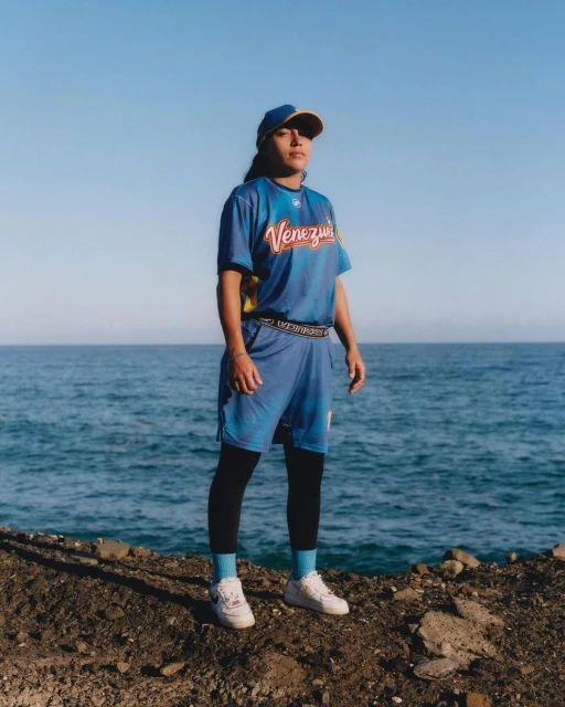 Woman in blue baseball uniform stands on rocky ground near the ocean on a clear day, wearing a cap and athletic shoes.