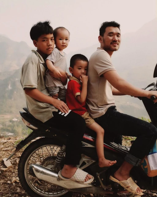 A man drives a moped on a rural road with three children riding with him. Mountainous background and soft natural lighting.