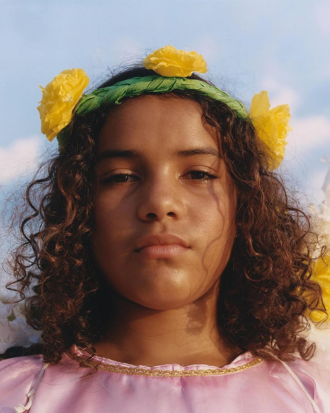 Portrait of a child with curly hair wearing a flower crown and pink top against a blue sky background, shot in medium close-up.