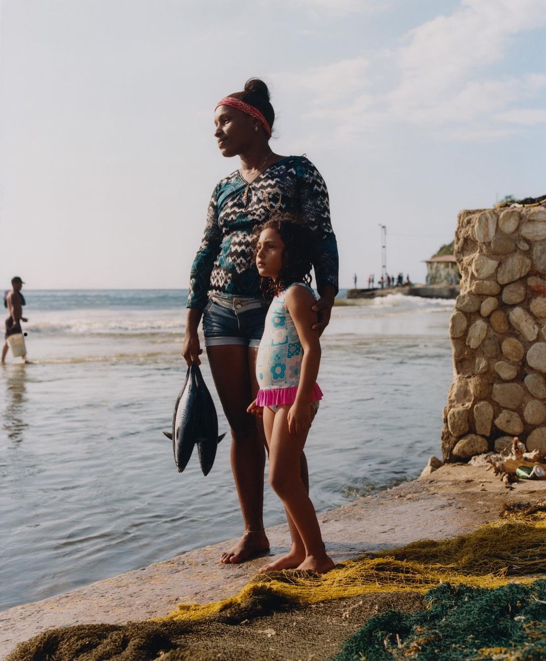 Woman and child stand on a concrete pier near the ocean with harvested fish in hand on a sunny day in a documentary style.