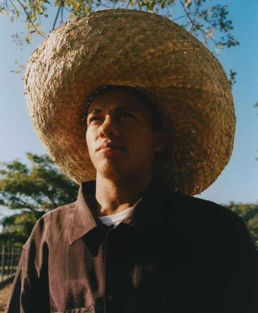 Portrait of a young man wearing a straw hat in an outdoor setting with trees and blue sky, capturing a sunny and casual atmosphere.