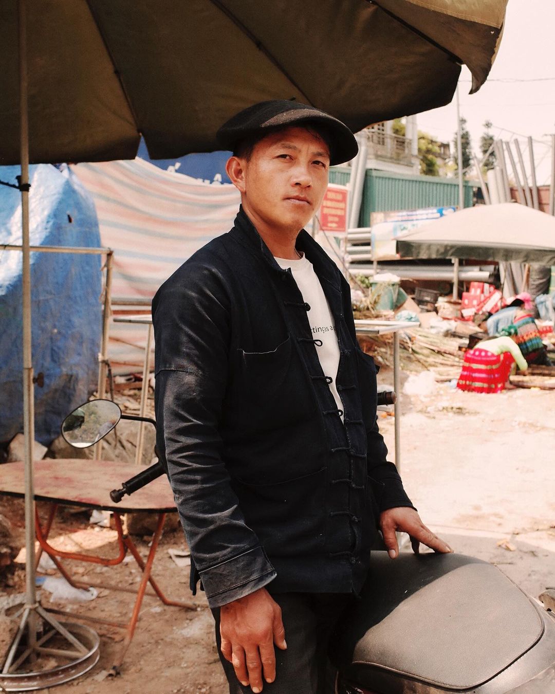 A middle-aged man with a cap stands near a motorcycle under an umbrella, wearing a dark jacket and light shirt, in an outdoor setting.