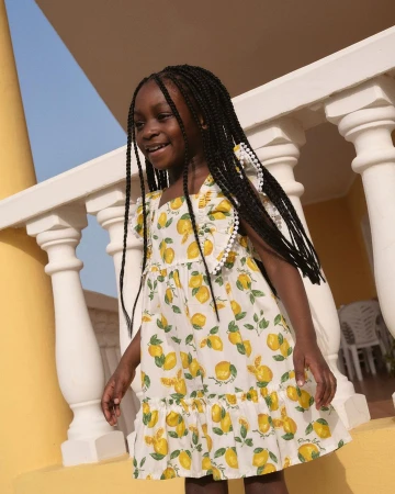 A cheerful young black girl smiles outdoors, wearing a summer dress with a lemon print and white hair accessories with a sky background.