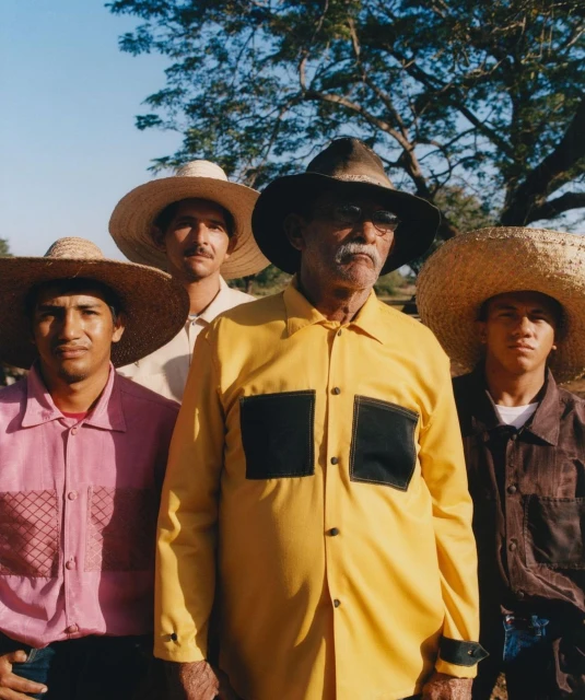 Group of four men in straw hats and colorful shirts pose outdoors with a tree visible in the background against a bright blue sky.
