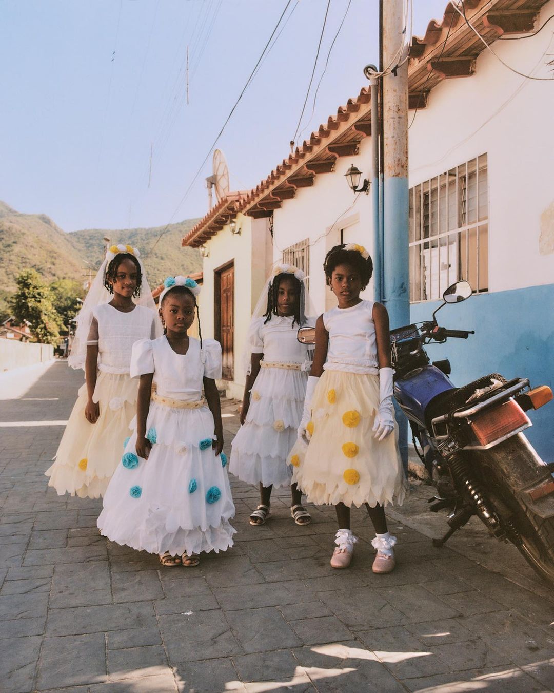 Four young girls stand in a row on a cobblestone street in decorative white dresses and veils, with a motorcycle parked nearby.