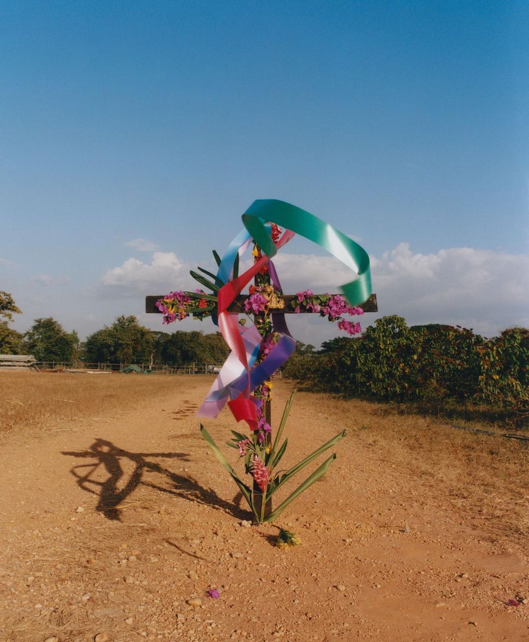 A cross decorated with flowers and ribbons stands in a dusty field under a clear blue sky, with distant trees completing the scene.
