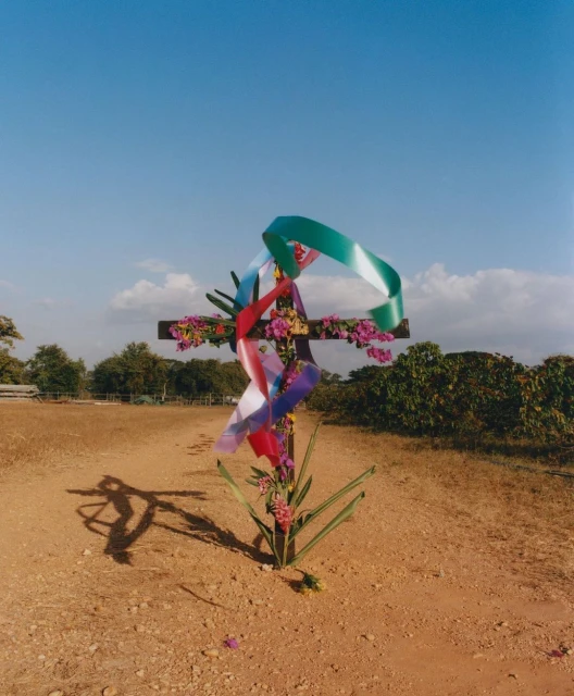 A cross decorated with flowers and ribbons stands in a dusty field under a clear blue sky, with distant trees completing the scene.