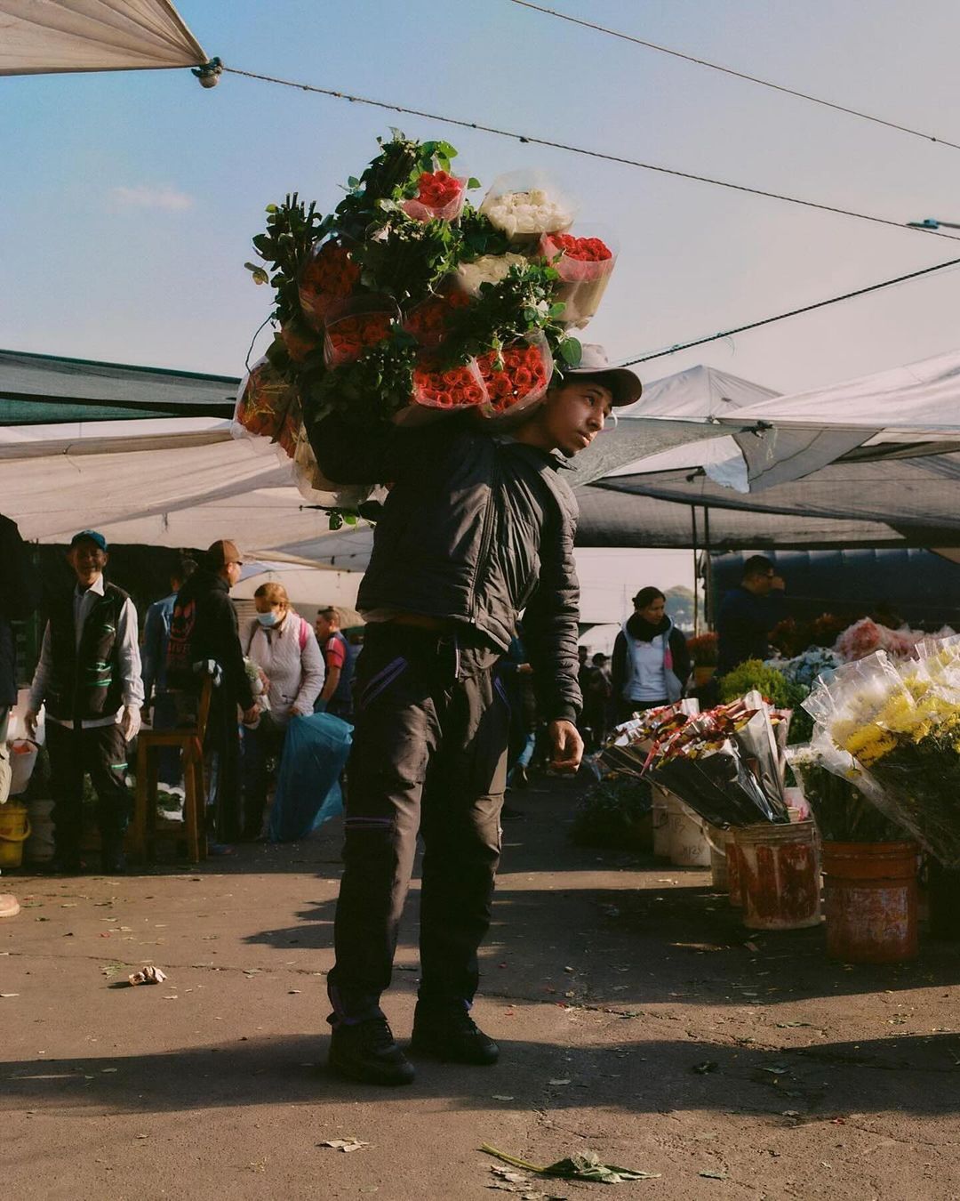 A person carrying bouquets of red and white roses over their shoulder at an outdoor market under tent canopies on a sunny day.