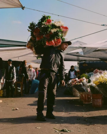 A person carrying bouquets of red and white roses over their shoulder at an outdoor market under tent canopies on a sunny day.