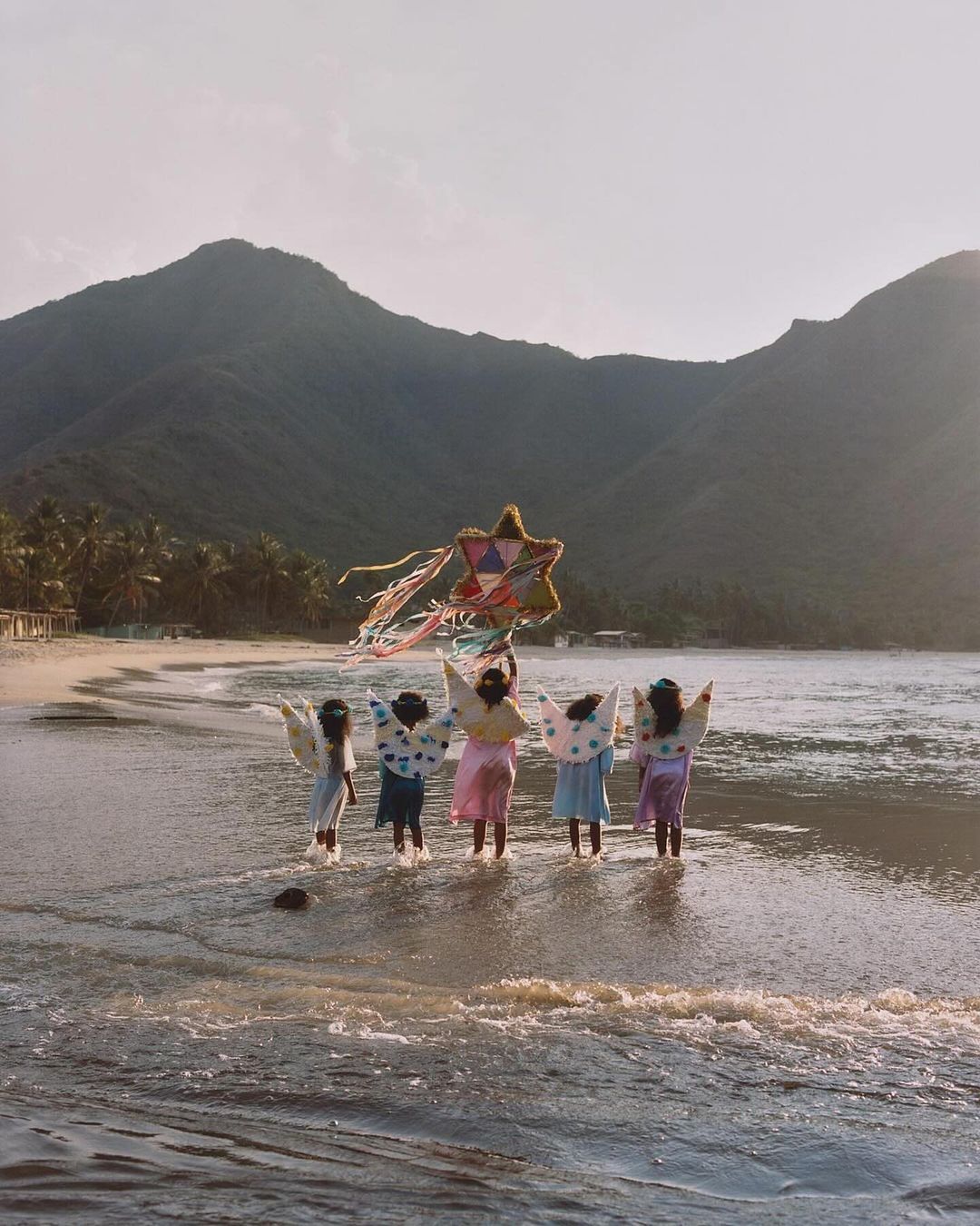 Group of children in angel costumes playing with a star kite on a sandy beach near mountains, creating a whimsical, joyful scene.