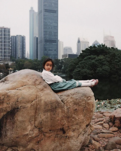 Young Asian girl in white blouse and green skirt sits on a large rock formation in a park with city buildings in the background.