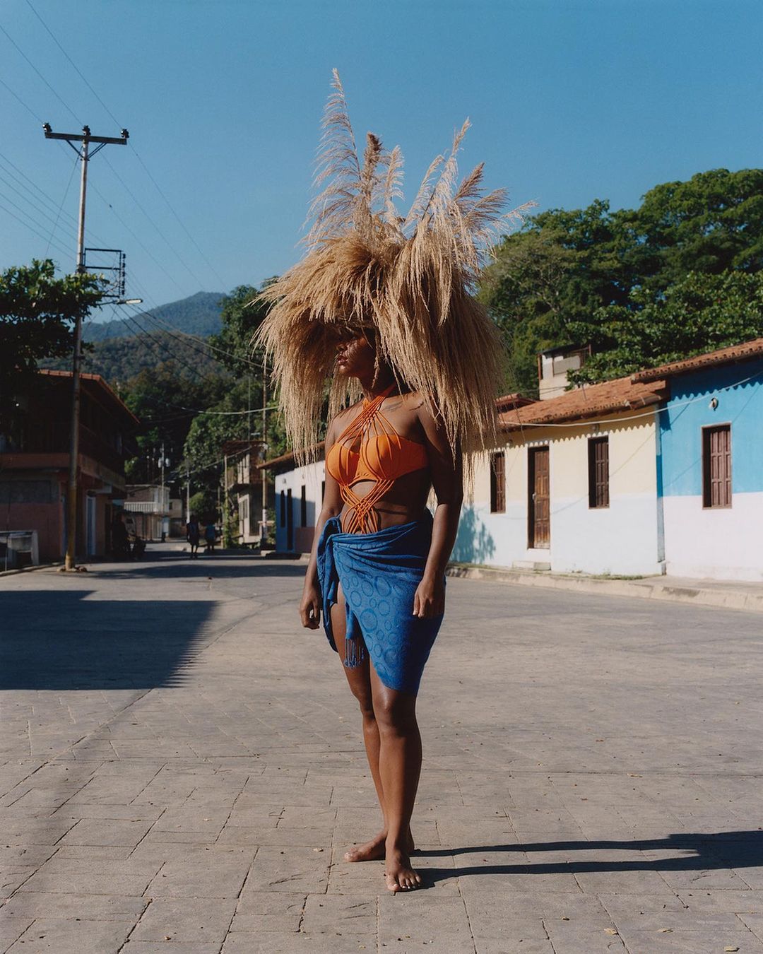 Woman in orange swimsuit and blue wrap stands on a paved road in a rural setting, wearing a large headpiece made of dried grass.
