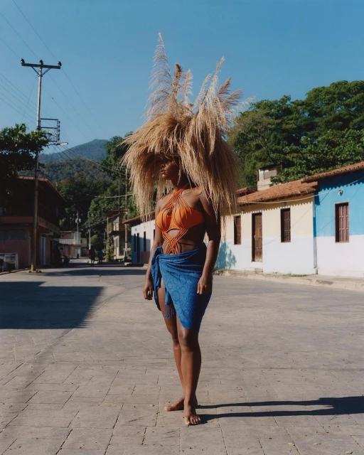 Woman in orange swimsuit and blue wrap stands on a paved road in a rural setting, wearing a large headpiece made of dried grass.