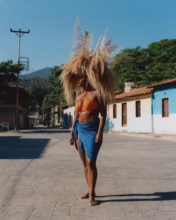 Woman in orange swimsuit and blue wrap stands on a paved road in a rural setting, wearing a large headpiece made of dried grass.