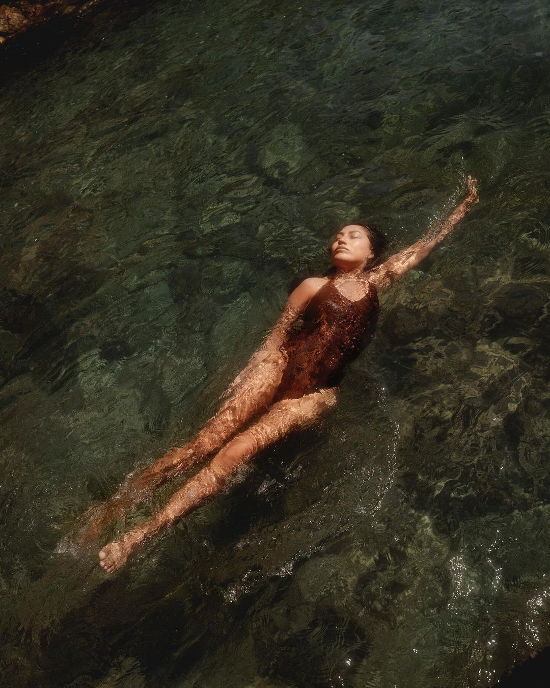 Woman in swimsuit floating on her back in the shallow clear water under the sun, eyes closed, one arm extended overhead, carefree.