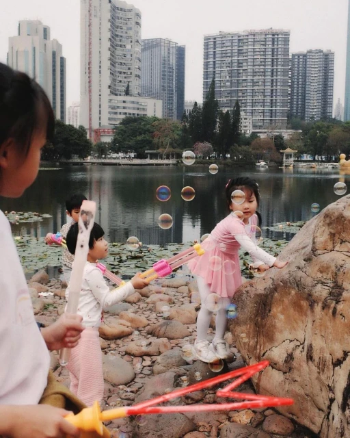 A group of young children play with bubble wands near a lake in an urban park, with city buildings in the background on an overcast day.