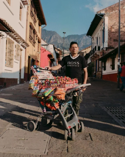 Adult male vendor stands behind a stroller converted to a candy cart on a city street with colorful buildings and mountains in the distance.