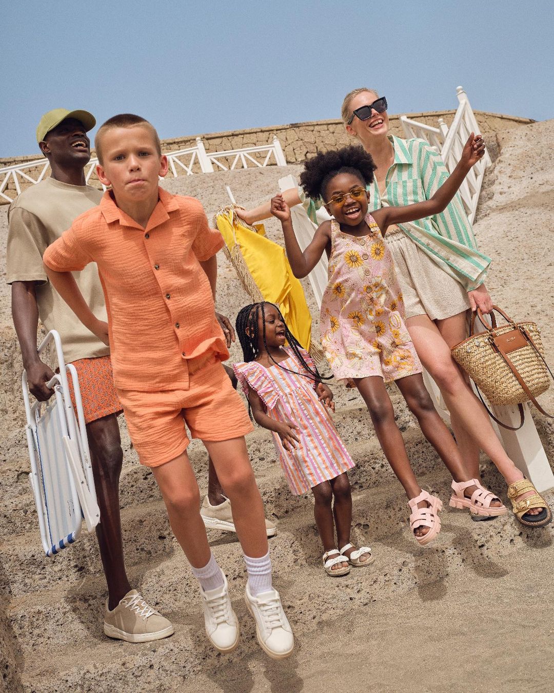 Group of adults and children in fashionable summer clothing posing on a beach dune with a straw bag and blue sky in the background.