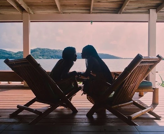 Two silhouetted figures sit in bamboo chairs on a wooden deck overlooking a tranquil lake framed by distant green hills.