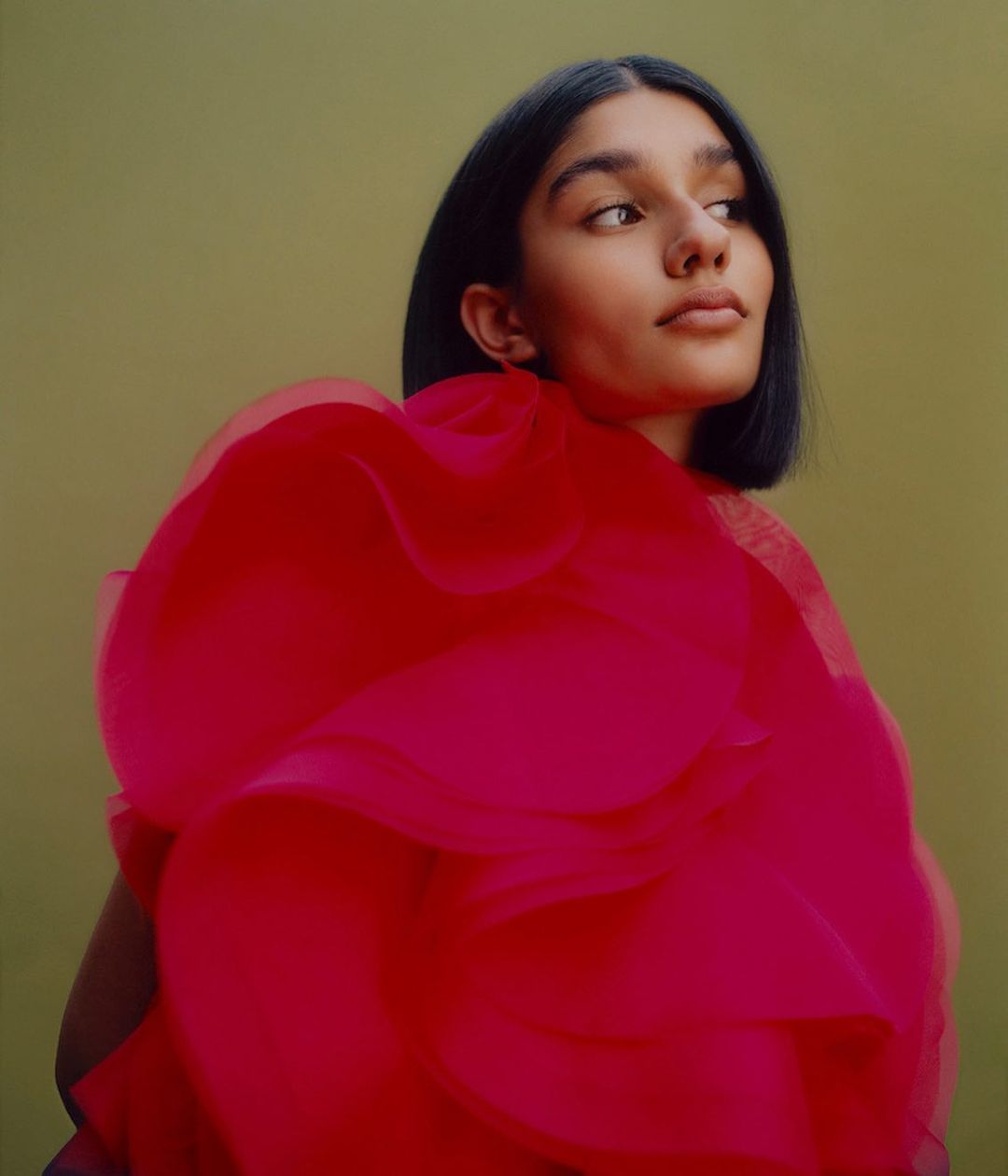 Portrait of a woman with a bob haircut, wearing a bright pink ruffled dress against a soft green background in a studio setting.