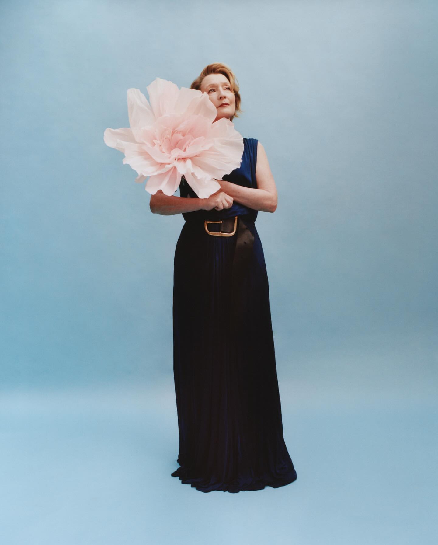 A woman poses in a navy dress with an oversized flower, standing against a light blue backdrop in a studio setting.