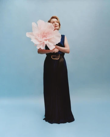A woman poses in a navy dress with an oversized flower, standing against a light blue backdrop in a studio setting.